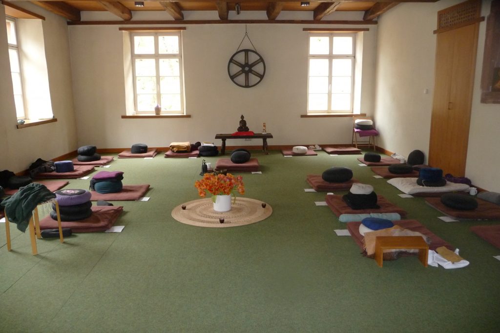 Bild zeigt einen ruhigen Meditationsraum mit einem großen braunen Ras in der Mitte, darunter ein Altar mit Buddhastatue